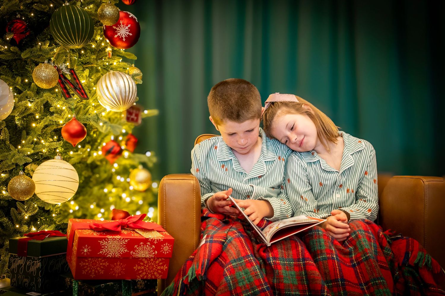 Two children in matching pajamas read a book together by a glowing Christmas tree with red ornaments.