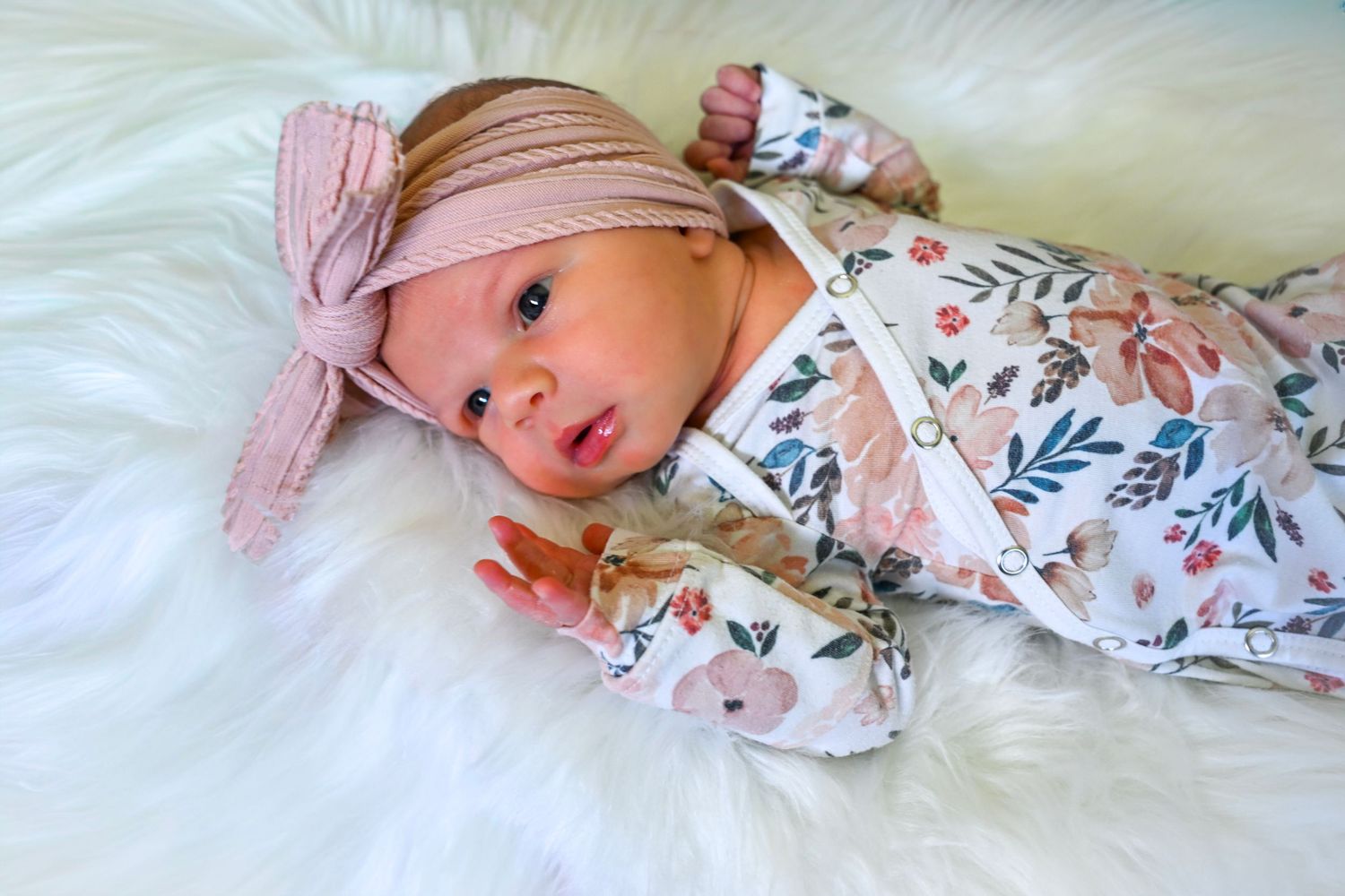 Newborn baby girl in floral onesie and pink bow headband lying on white fluffy blanket.