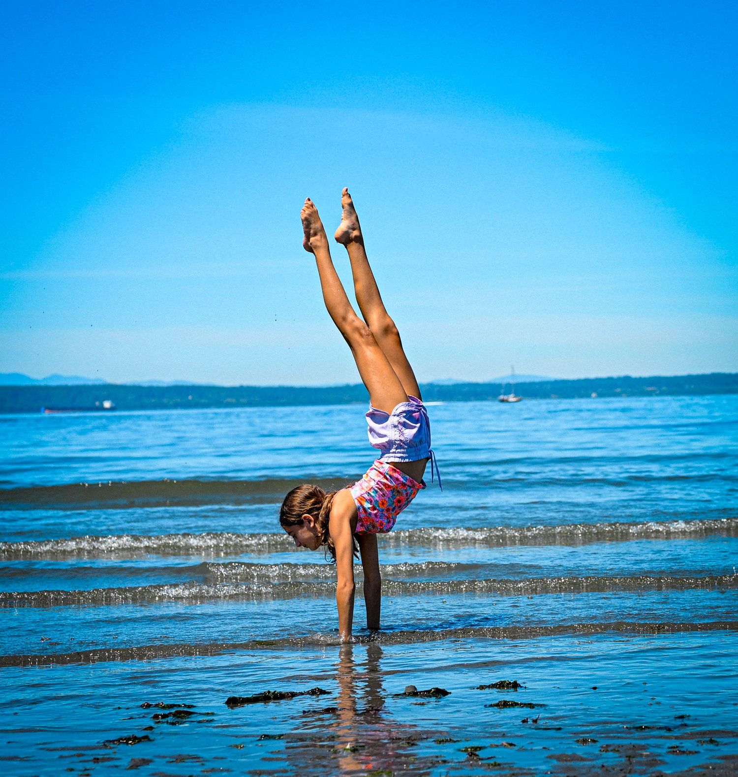 A young girl performs a perfect handstand on a sunny beach with calm blue ocean waves in the background.