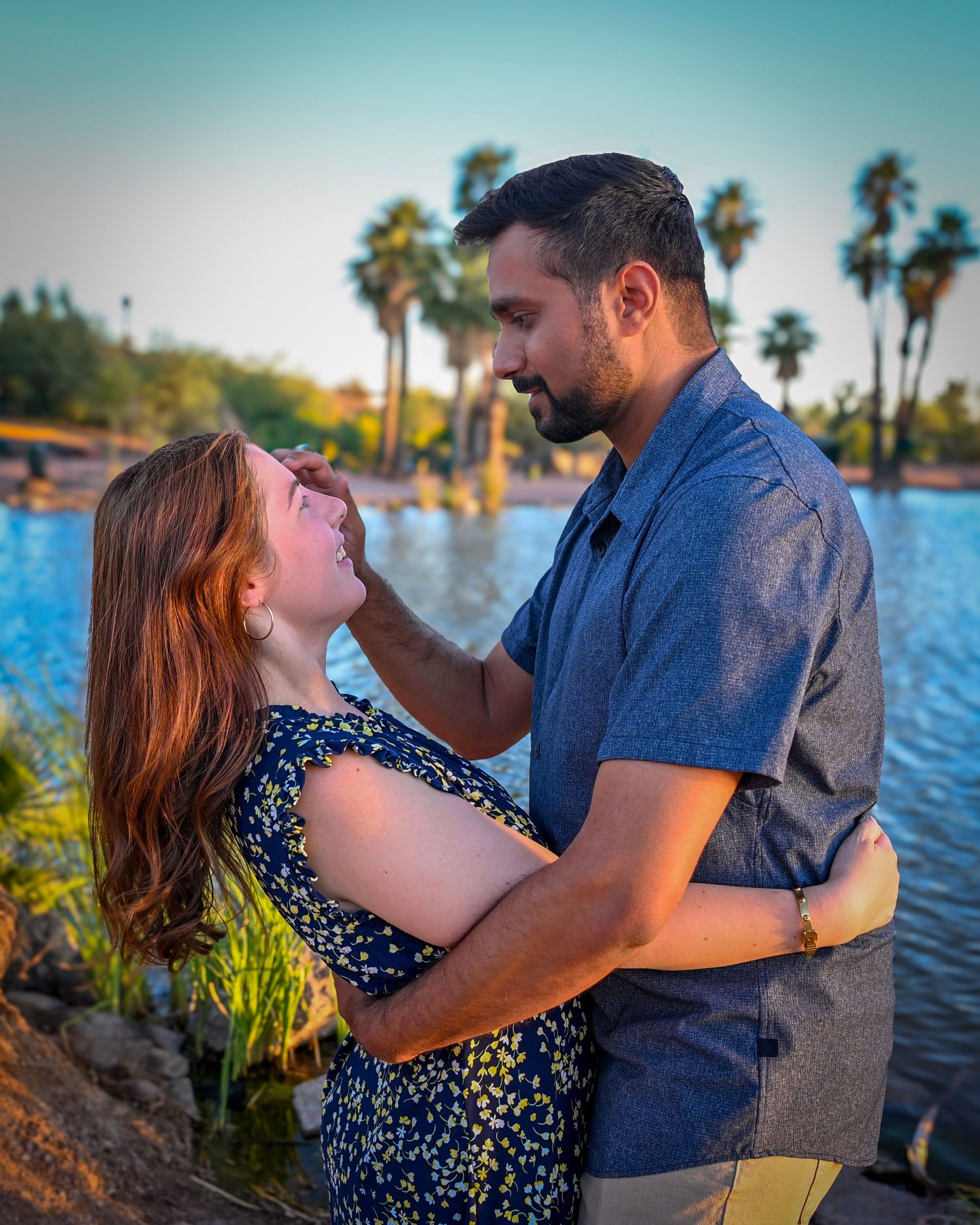 A couple shares a tender moment by a palm-lined lake at sunset, the man gently touching the woman's face.