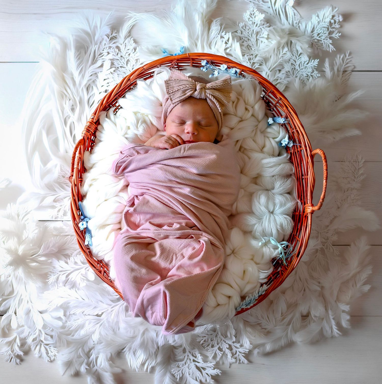 Newborn baby swaddled in pink wrap with bow headband sleeping in wicker basket on white snowflake backdrop.