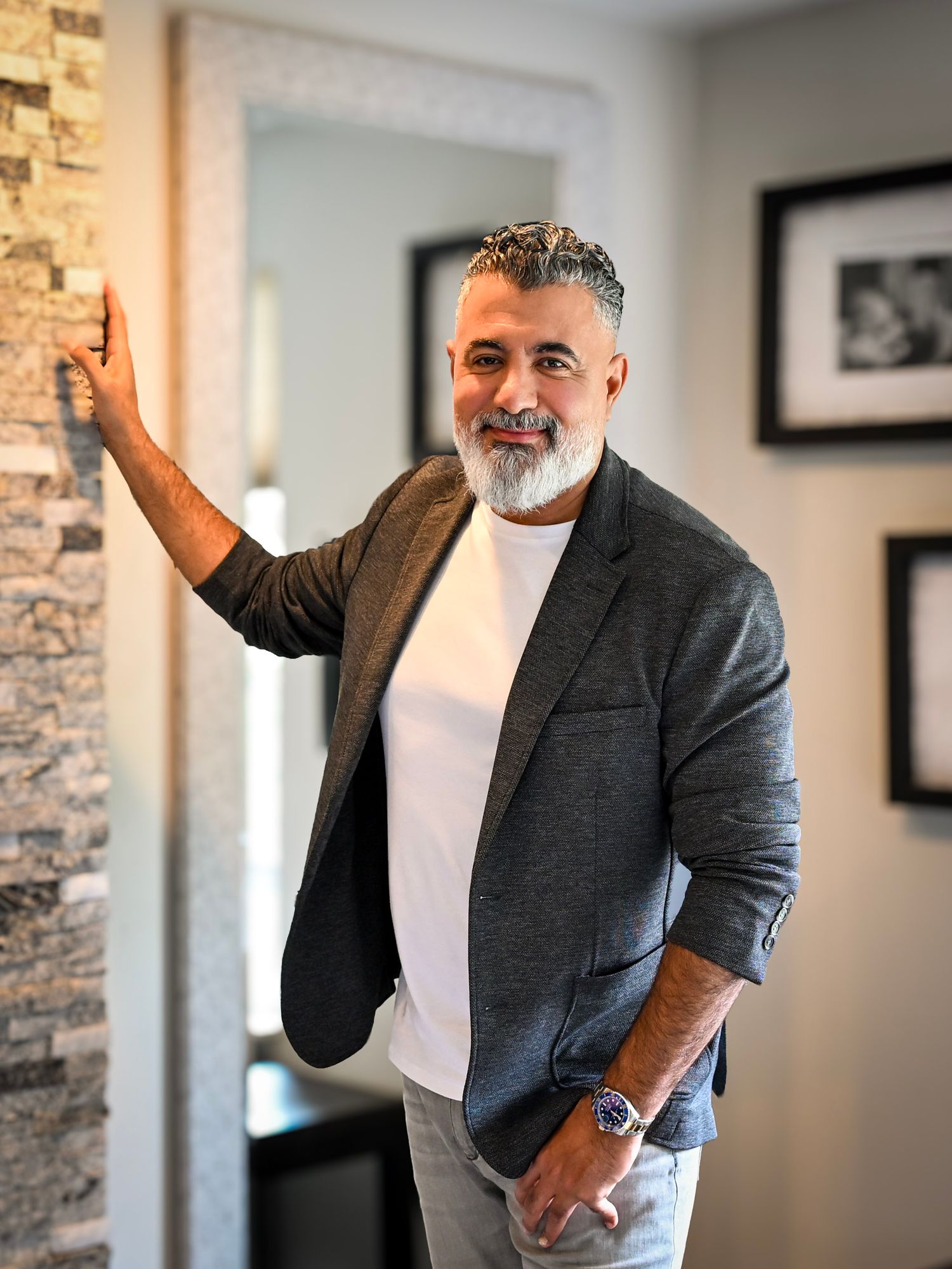 A well-dressed mature person in a blazer and white t-shirt poses casually against a wall with framed artwork in the background.