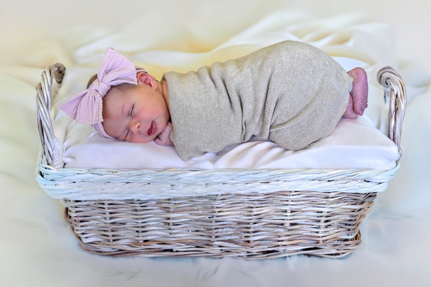 Sleeping newborn baby girl swaddled in gray wrap with pink bow in a white wicker basket.