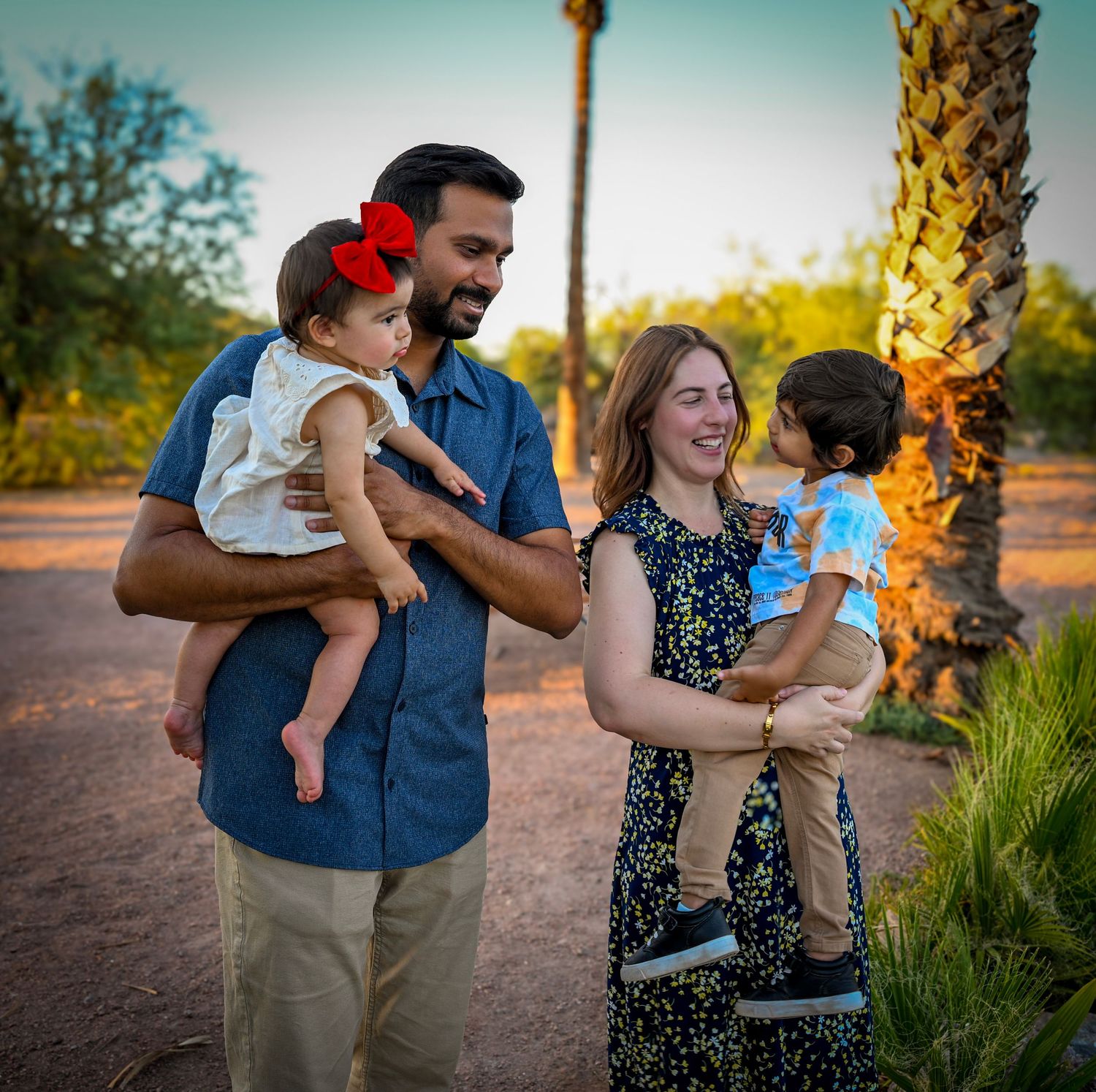 A happy family of four poses outdoors near a palm tree at sunset in a desert garden setting.