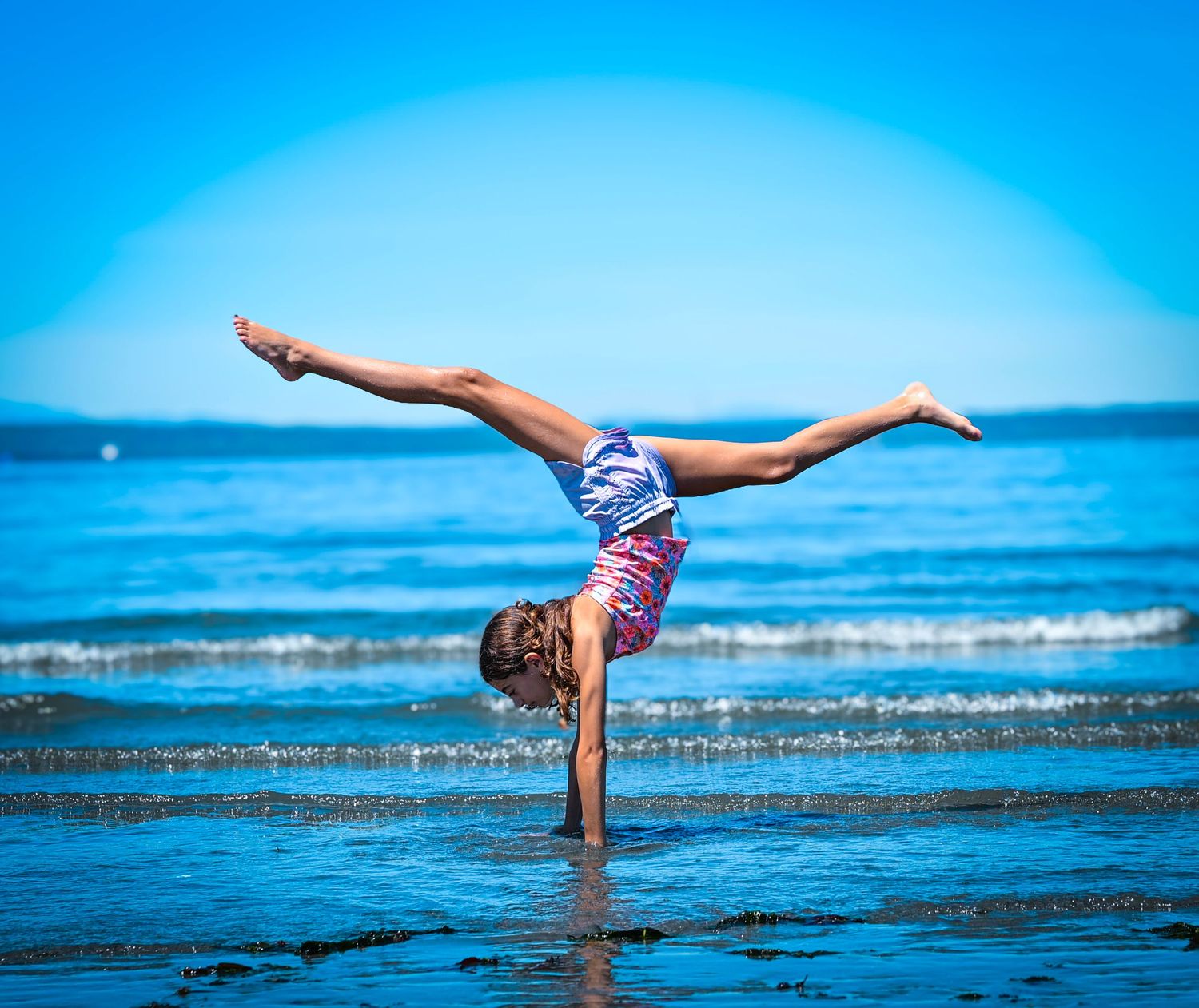 A gymnast performs a one-handed handstand on a beach with legs split, wearing a colorful leotard against a blue ocean backdrop.