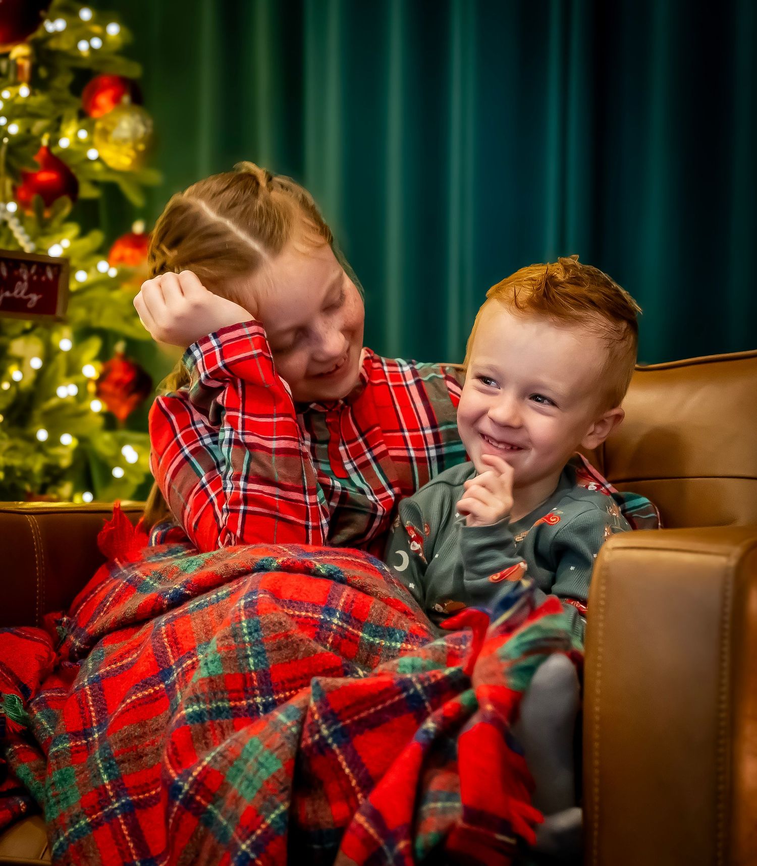 Two children in festive plaid sharing a warm moment by a decorated Christmas tree.