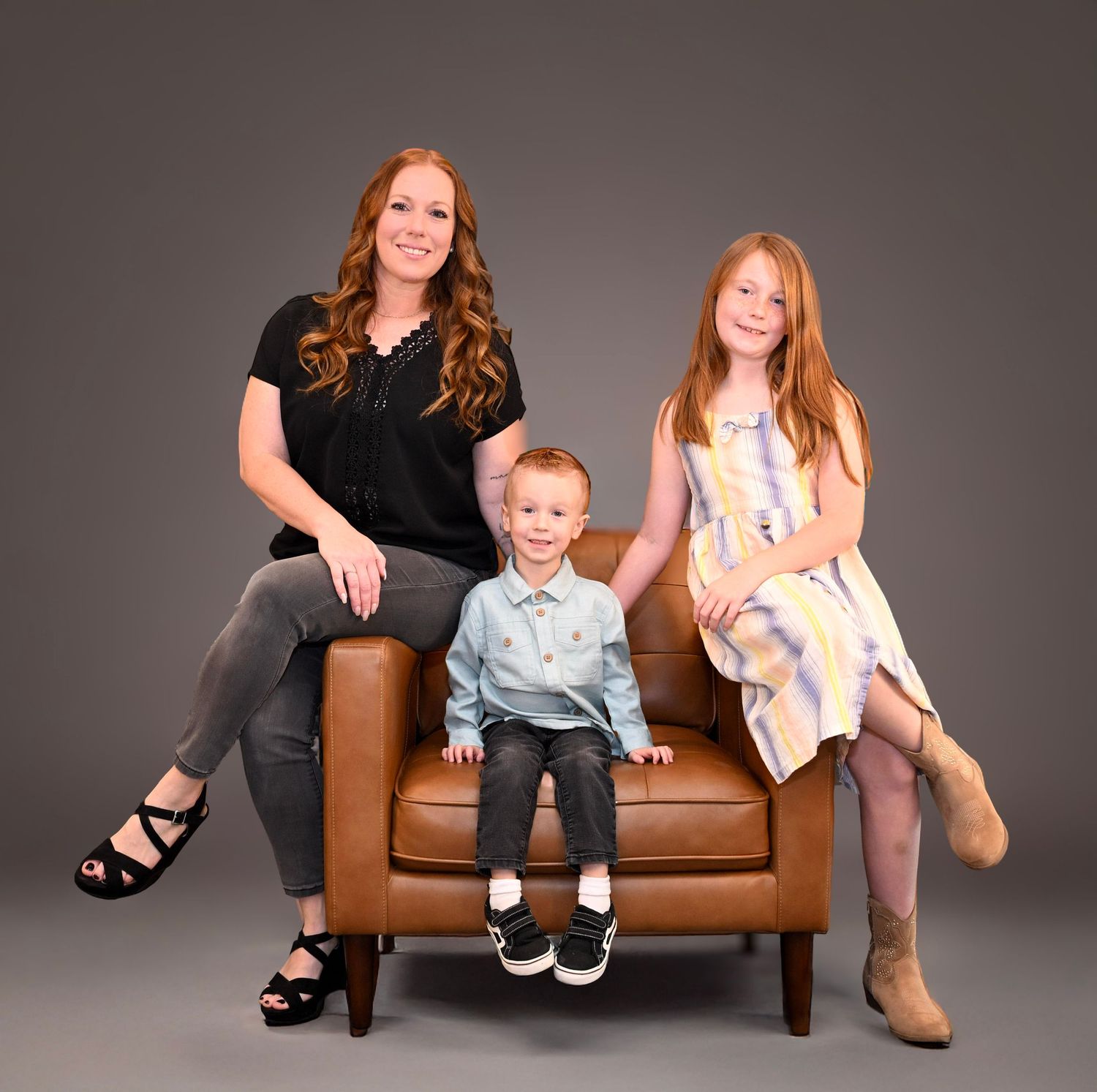 A family group sits together on a brown leather bench against a dark gray studio backdrop.