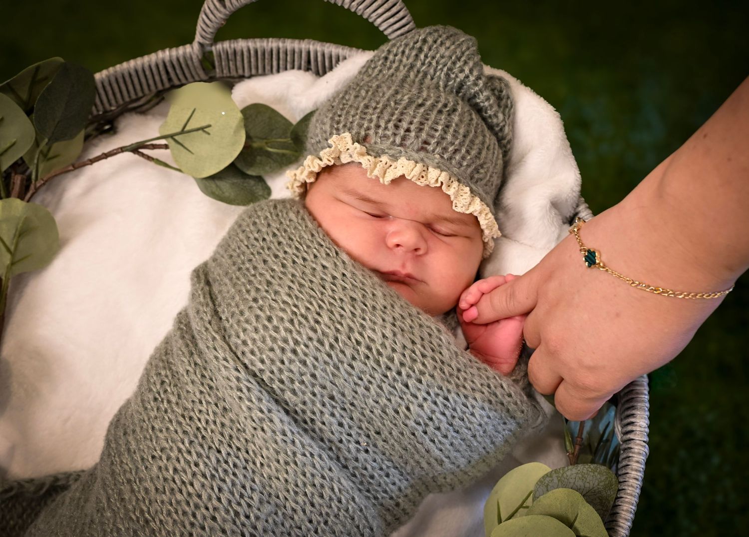 Newborn baby swaddled in gray knit wrap and hat, sleeping peacefully in a basket while an adult holds their tiny hand.
