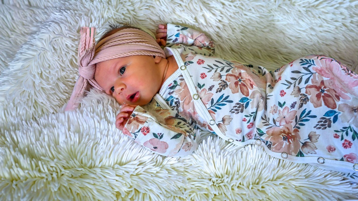 Newborn baby girl in floral outfit and pink headband resting on a white fluffy blanket.