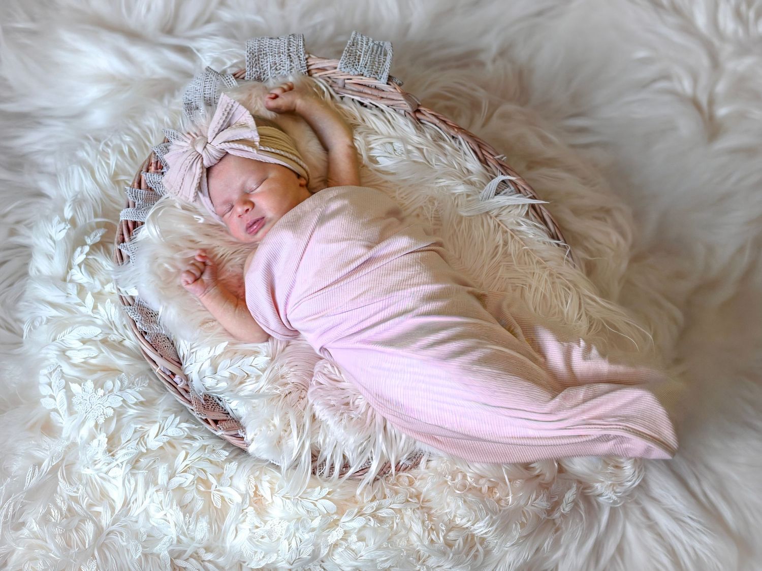 Newborn baby girl swaddled in soft pink wrap with gray bow headband sleeping in decorated wicker basket.