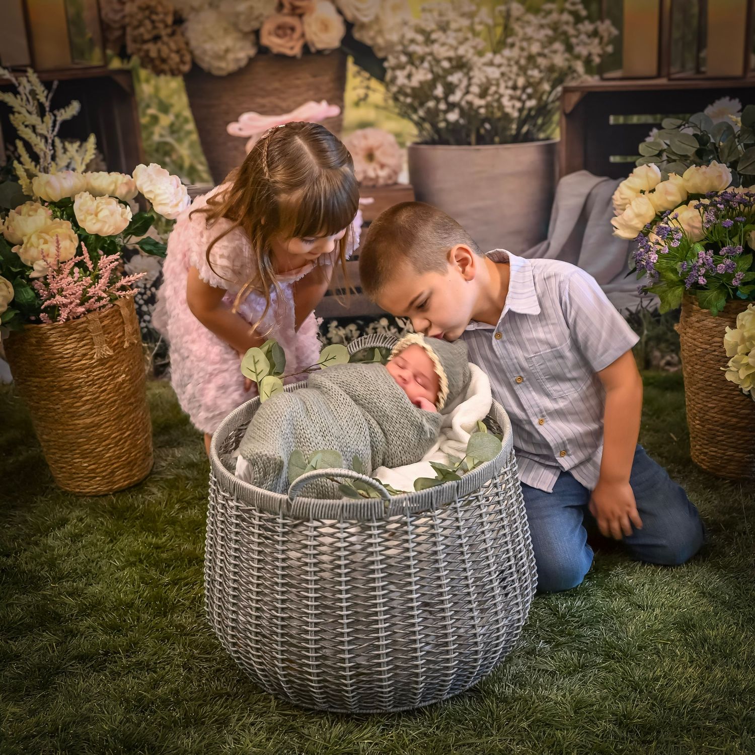 Two children lovingly gaze at a newborn baby in a wicker basket surrounded by flowers in a studio portrait.