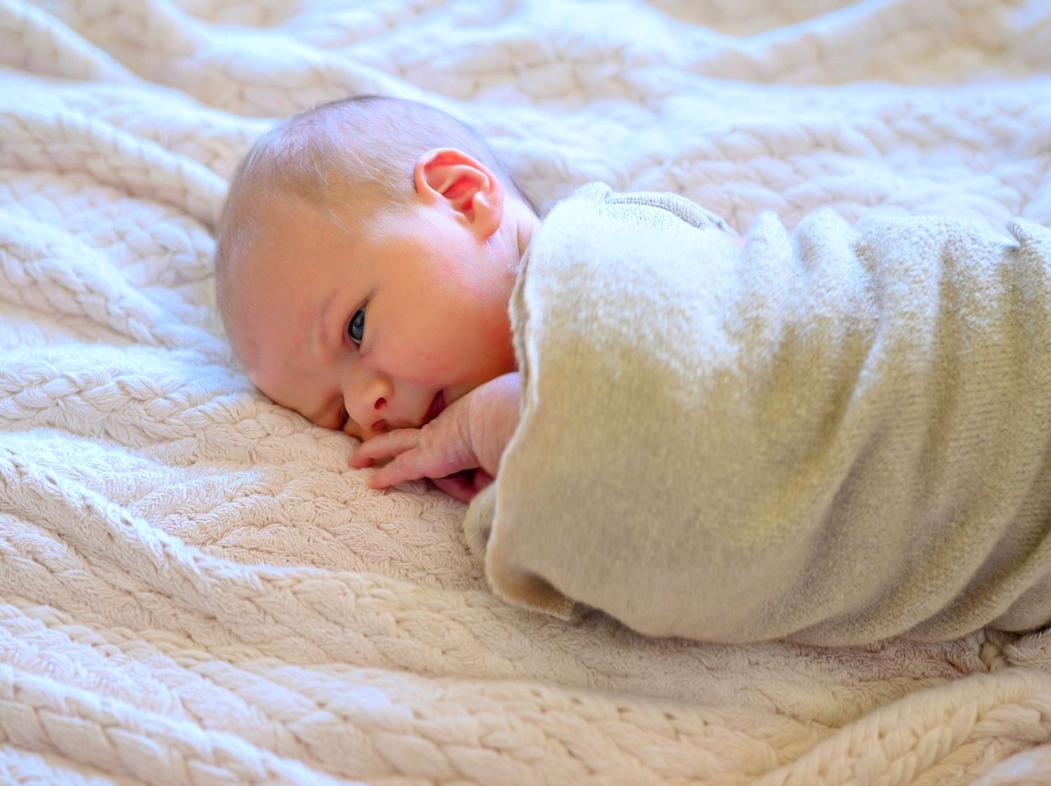 Newborn baby swaddled in a cream blanket lying on a quilted white surface.