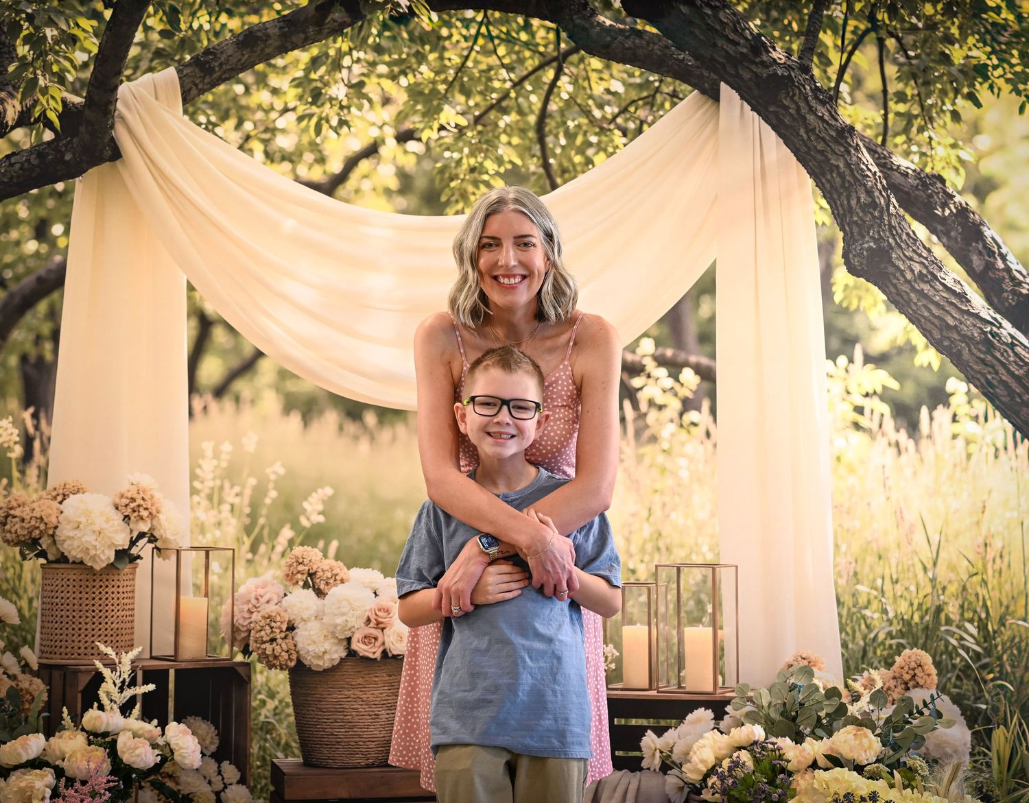 Woman and young boy with glasses pose under a floral arch with candles and blooms in a garden setting.