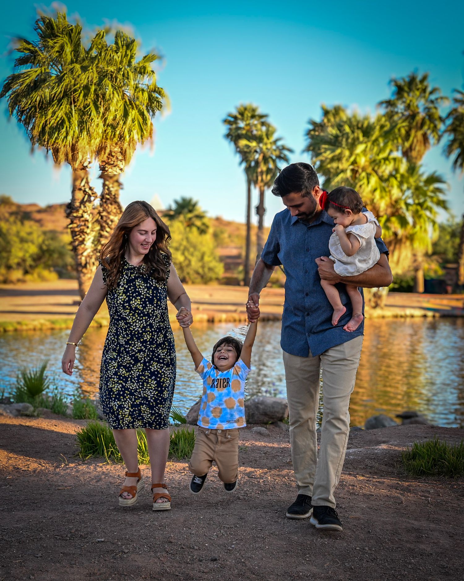 Family of four walking near a palm tree-lined lake, parents swinging toddler while dad holds baby.