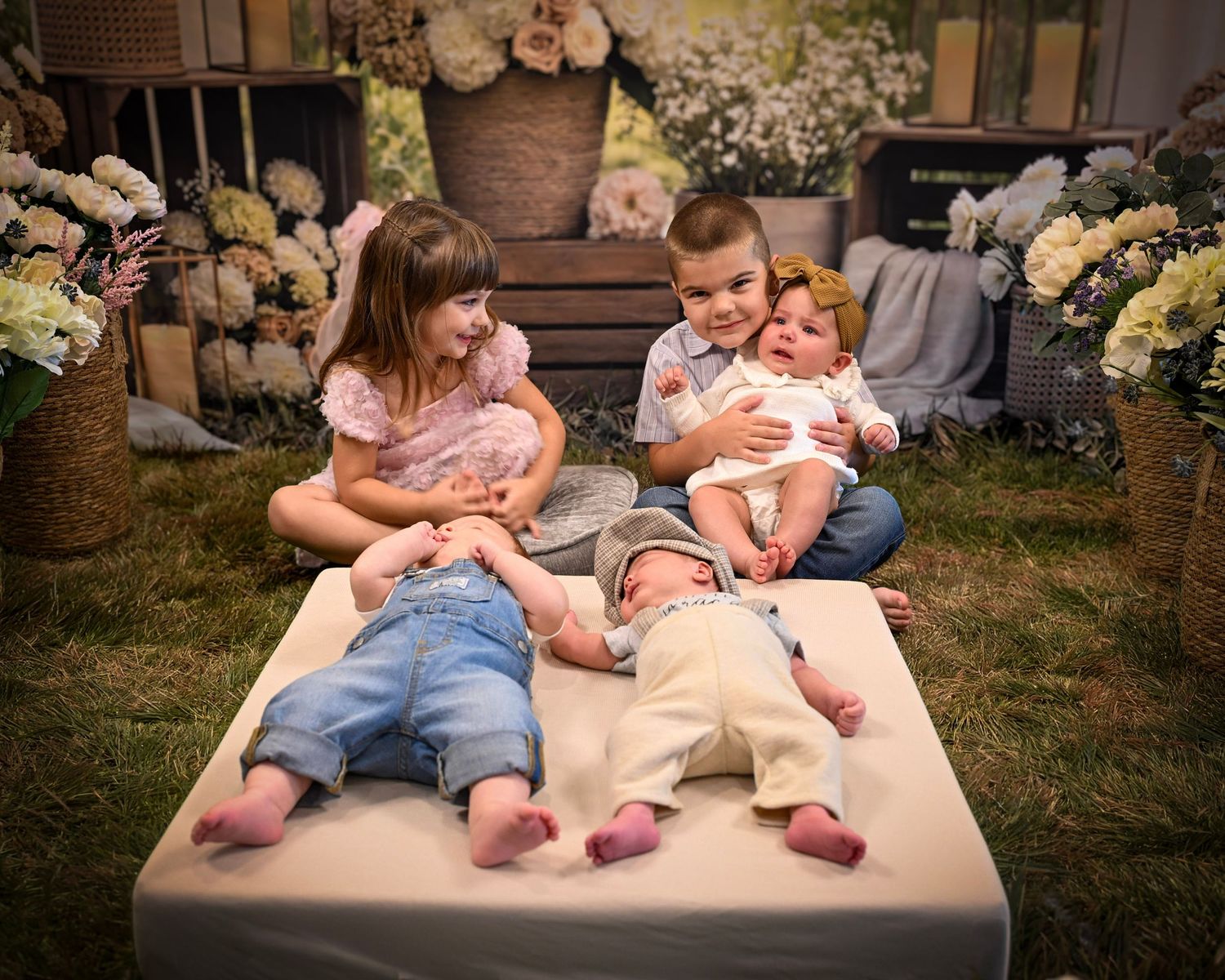 Two young children sit smiling with two newborn babies on a mat, surrounded by flowers in a charming garden photo shoot.