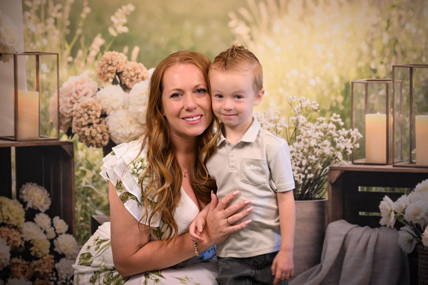 A smiling woman with long auburn hair poses with a young boy in a white shirt against a floral backdrop.