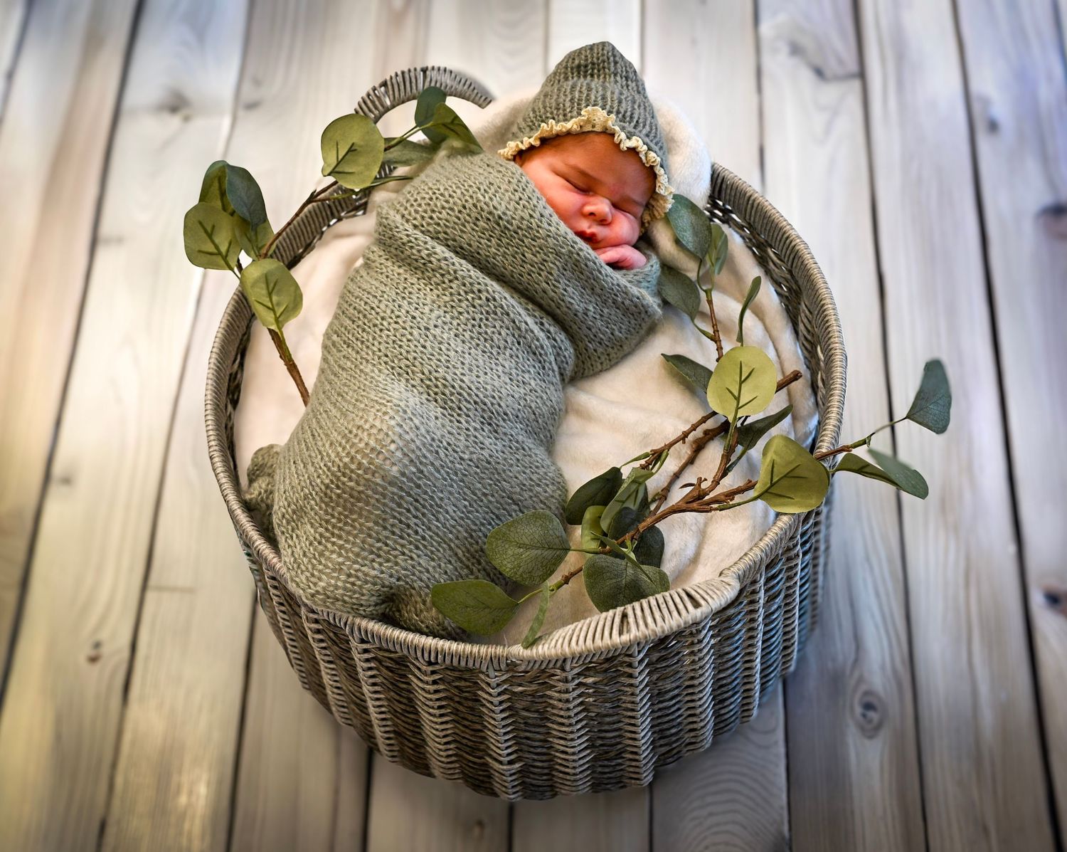 Sleeping newborn baby swaddled in gray knit wrap with matching bonnet nestled in wicker basket with eucalyptus.