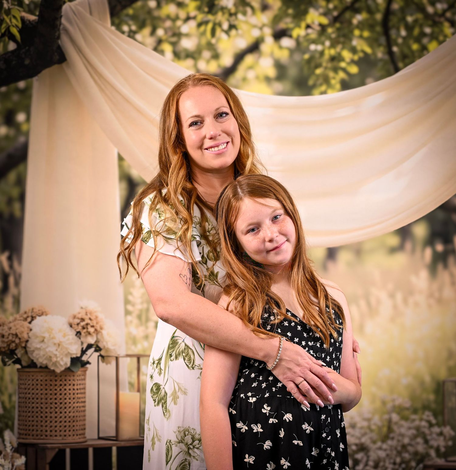A redheaded mother and her daughter share a warm embrace in a professional outdoor portrait with draped fabric backdrop.