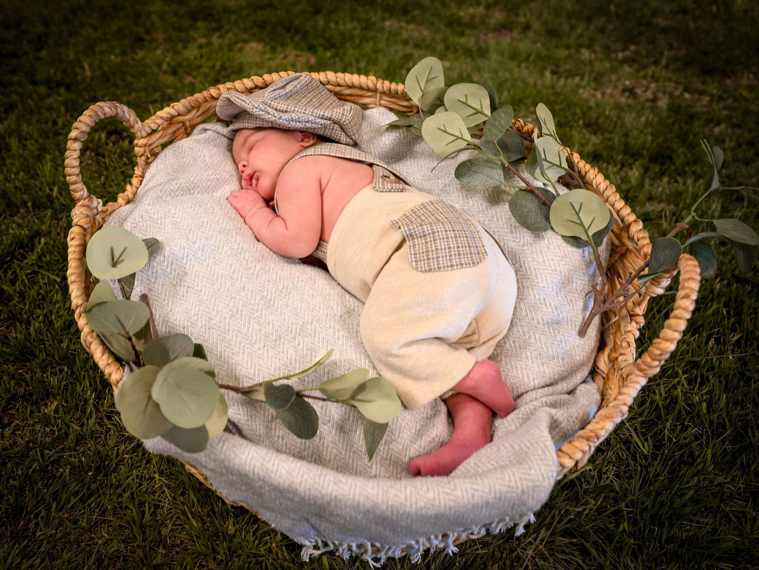 Newborn baby sleeping peacefully in a wicker basket with eucalyptus branches on a soft blanket outdoors.
