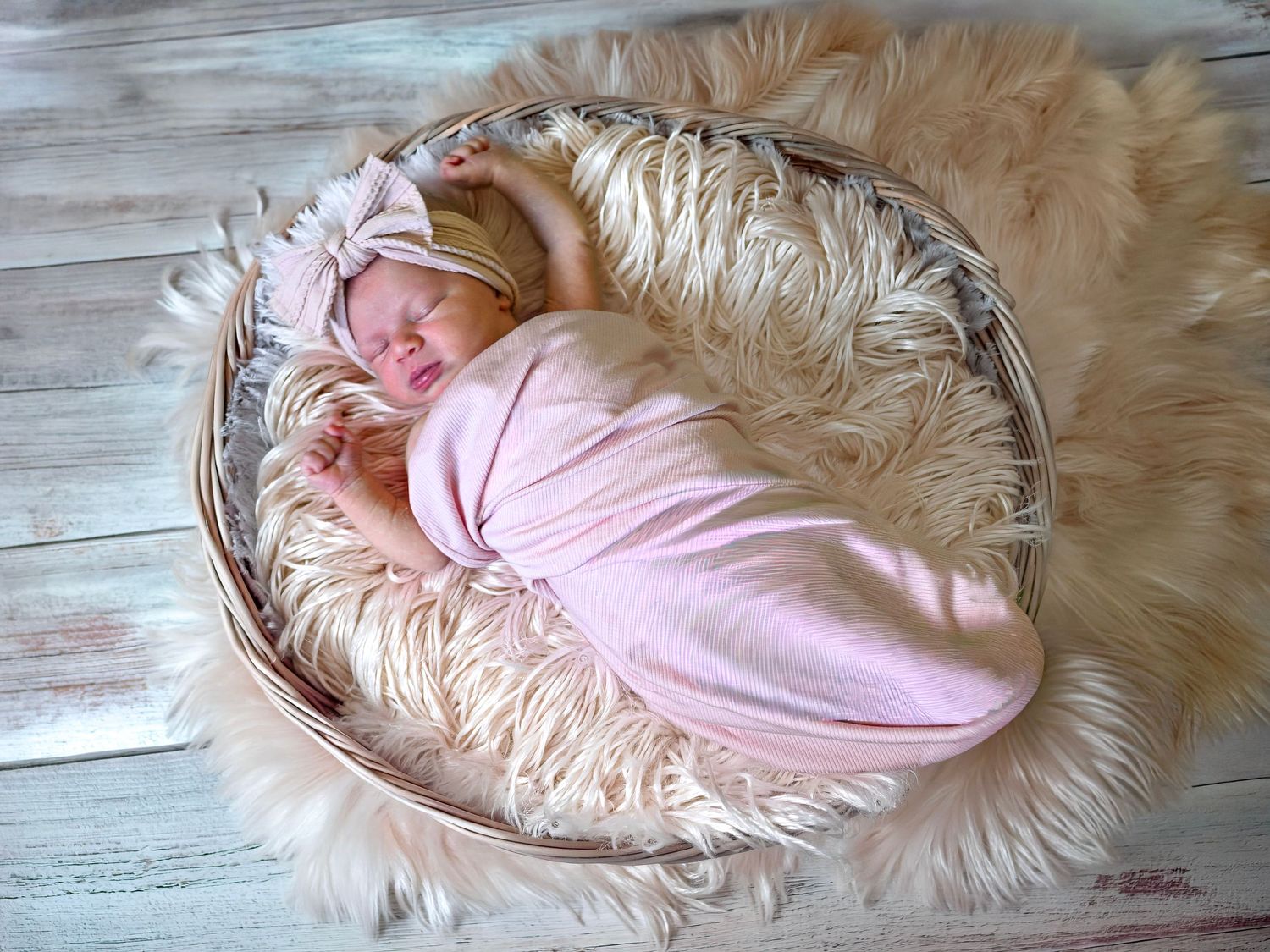 Newborn baby sleeping in wicker basket with pink swaddle and bow headband on fur.