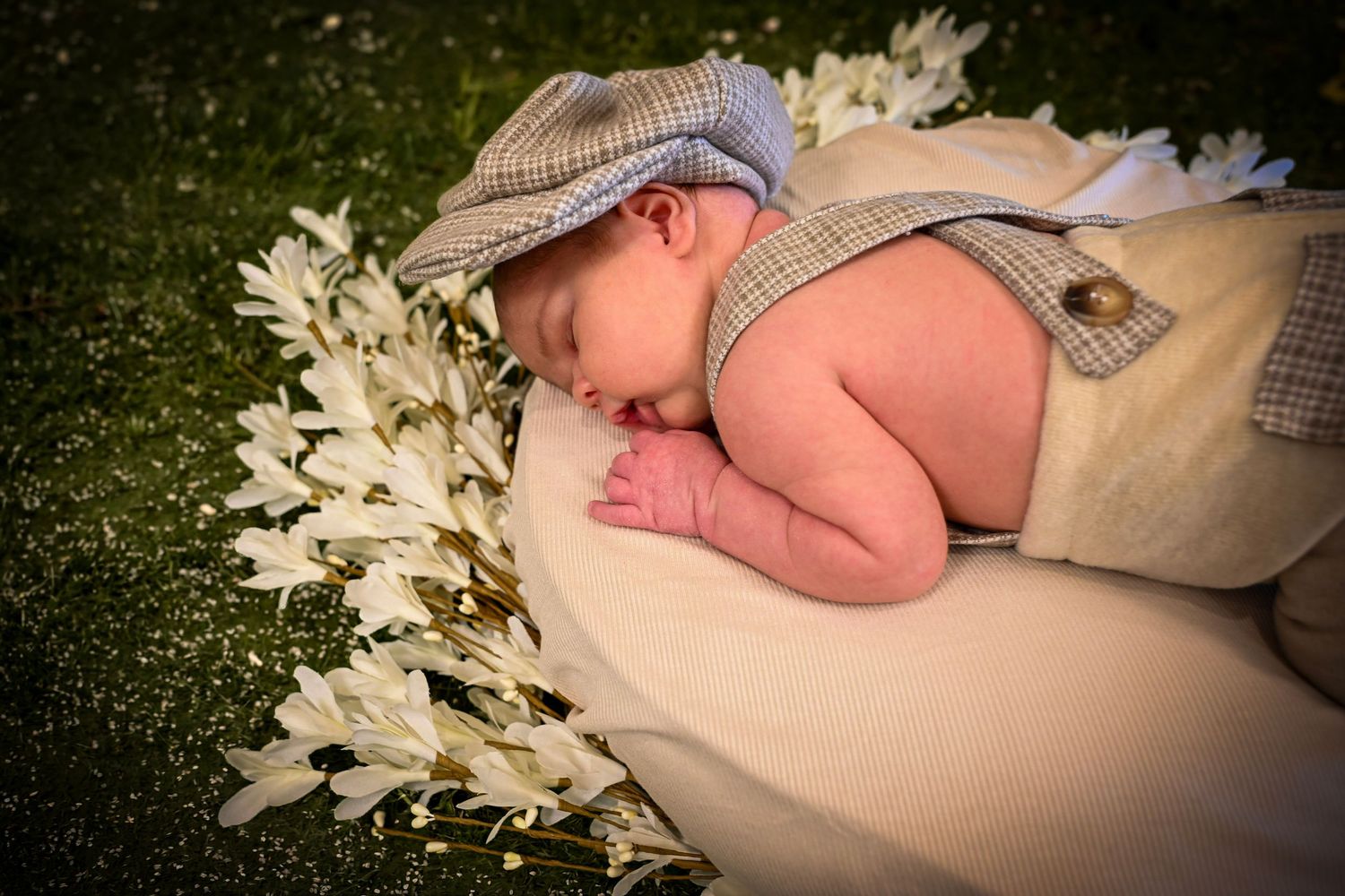 Newborn baby sleeping peacefully in a bowl wearing a plaid newsboy cap and suspenders, surrounded by white flowers outdoors.