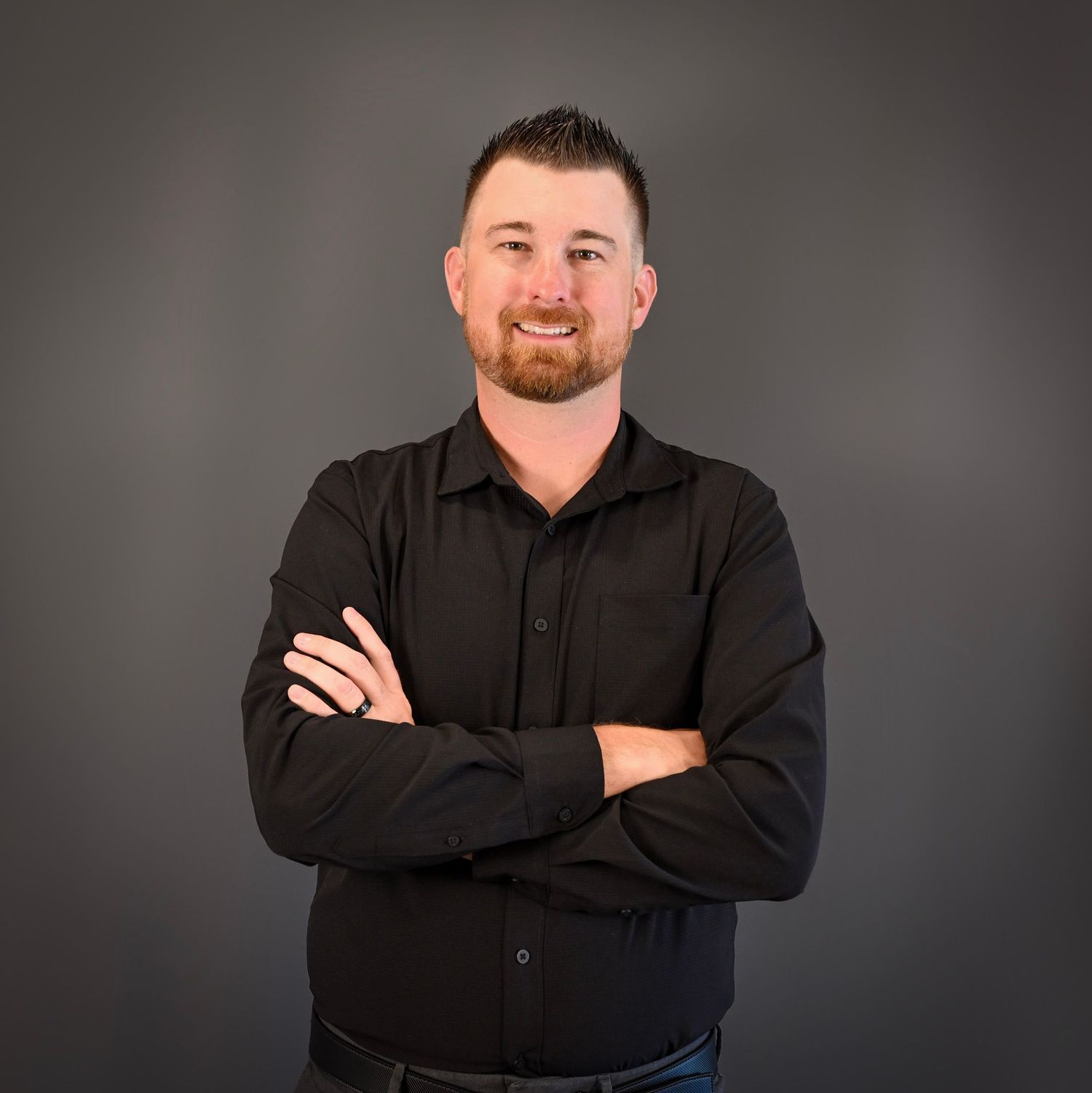 A smiling man with a beard in a black shirt stands with arms crossed against a gray background.
