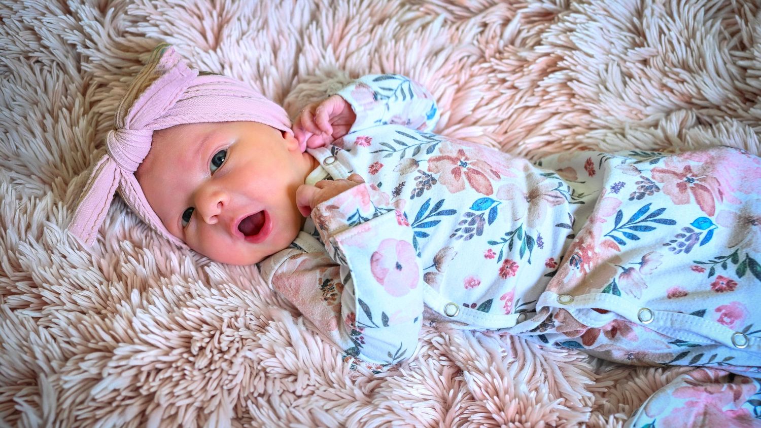Adorable newborn baby in floral outfit and pink bow hat lying on fluffy rug.