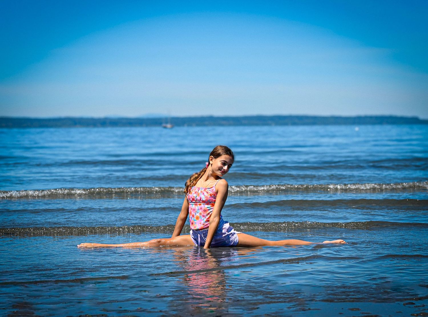 Young girl performing splits on a shallow ocean beach in a pink swimsuit on a sunny day.