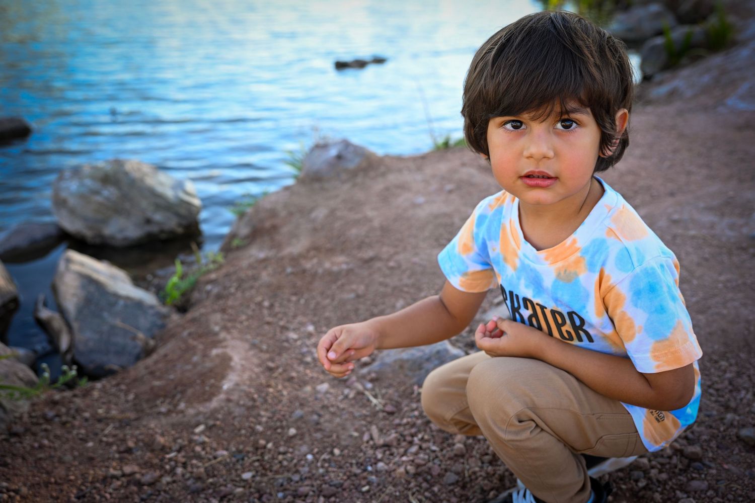 Young boy crouching near rocky shoreline wearing colorful tie-dye shirt, looking at camera by blue lake.