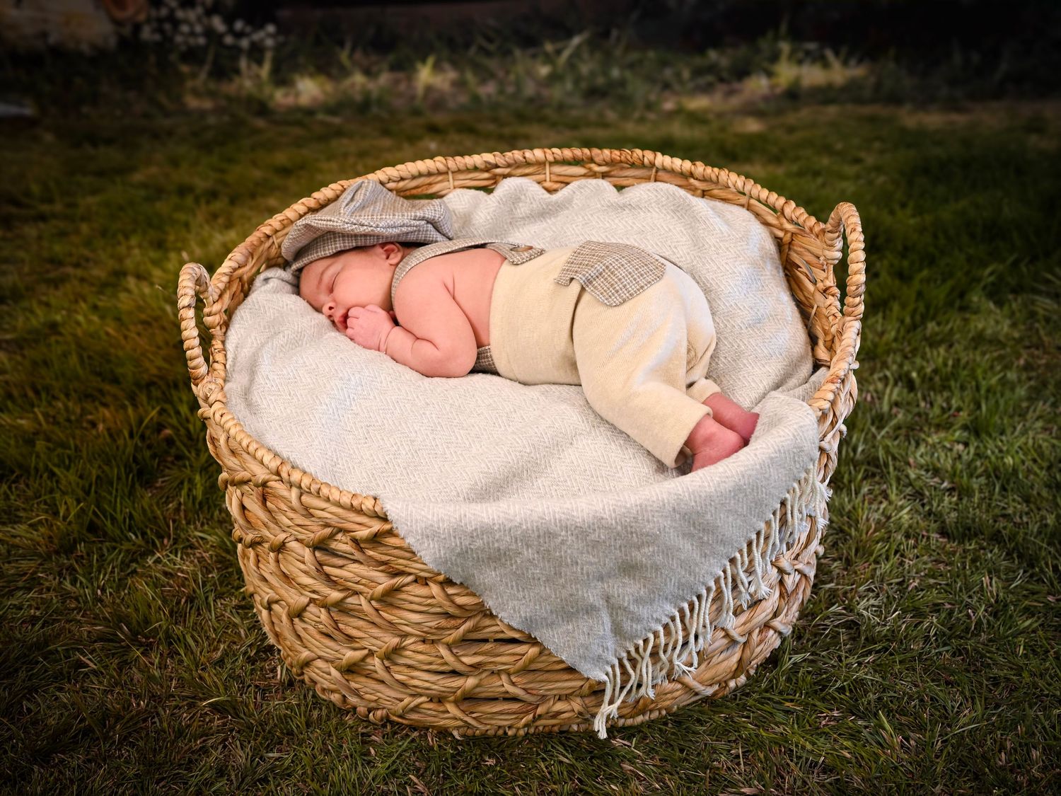 Newborn baby in cream outfit sleeping curled up in a large wicker basket on green grass outdoors.
