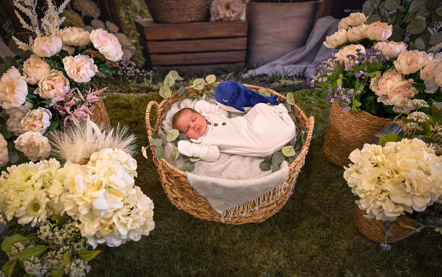 Newborn baby sleeping peacefully in a wicker basket surrounded by beautiful white and pink floral arrangements.
