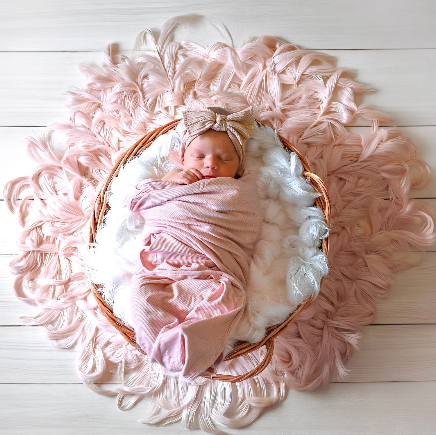 Sleeping newborn wrapped in pink swaddle with bow headband nestled in wicker basket on ruffled pink fabric mat.