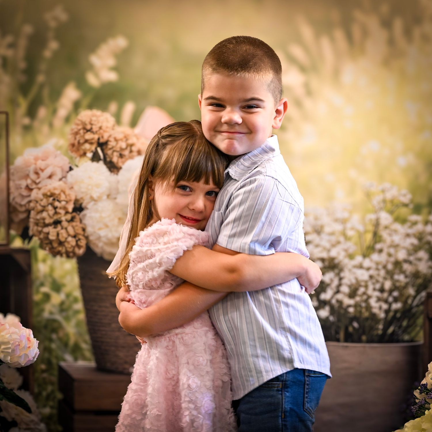 Two young children hugging and smiling in front of a floral backdrop, dressed in pink dress and blue shirt.