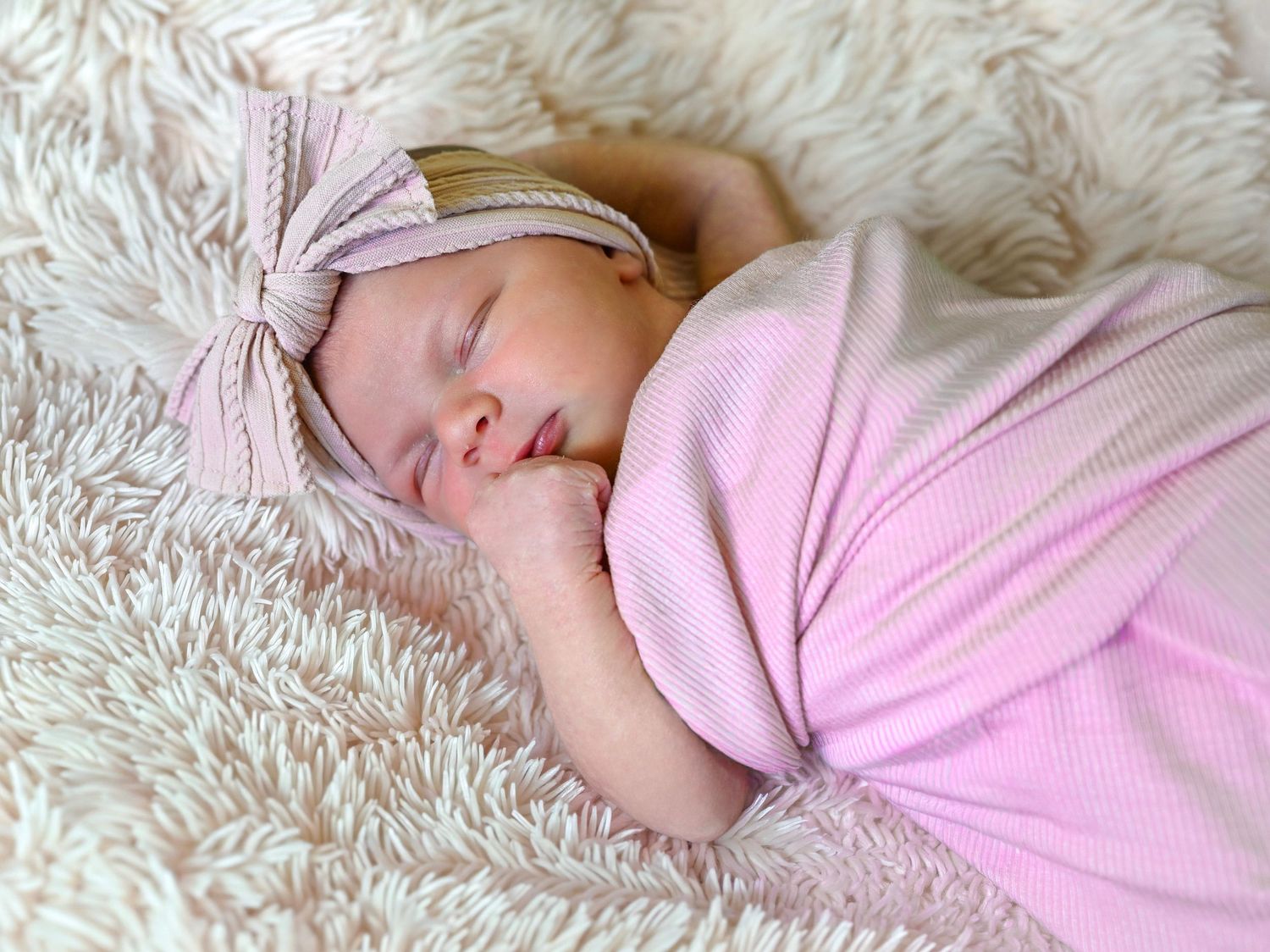 Newborn baby girl sleeping peacefully swaddled in pink blanket with bow headband on fluffy white background.