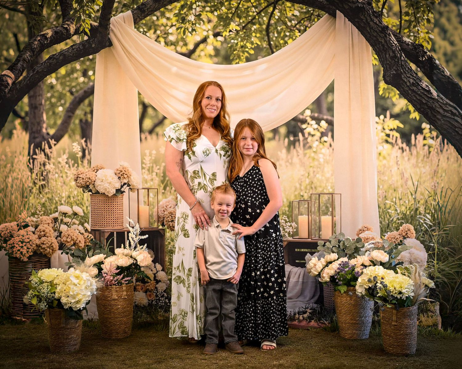 Woman and two children posing outdoors under draped fabric arch surrounded by wildflowers and hydrangeas.