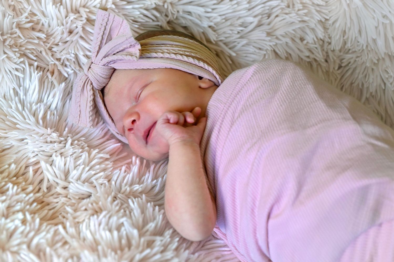 Newborn baby girl sleeping peacefully wrapped in pink swaddle with bow headband on fluffy white blanket.