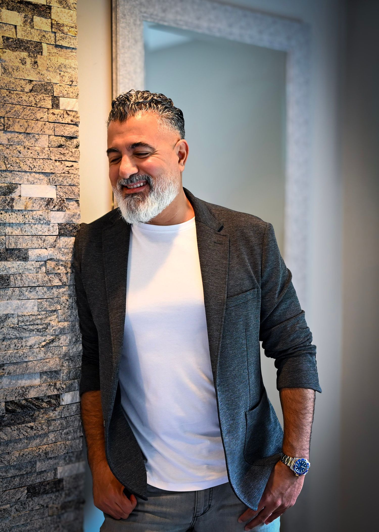 A person wearing a gray blazer and white t-shirt poses against a textured stone wall in a professional setting.