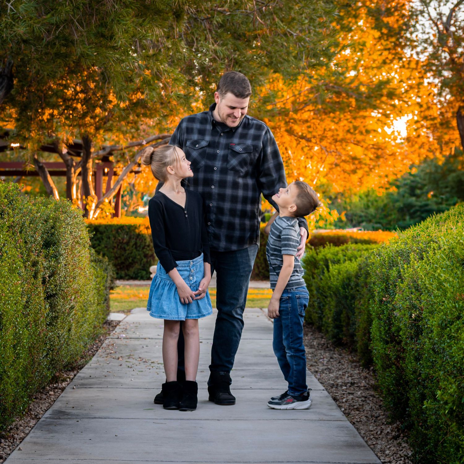 A family walks along a garden path surrounded by autumn foliage and hedges during golden hour.