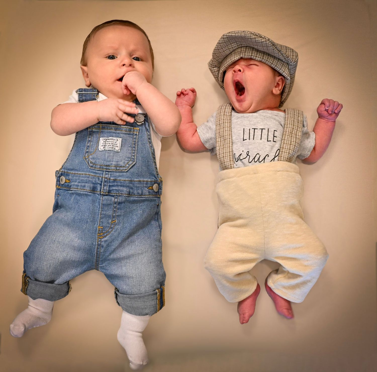 Two adorable babies lying side by side, one in denim overalls and one in a newsboy cap and Little Rascals outfit.