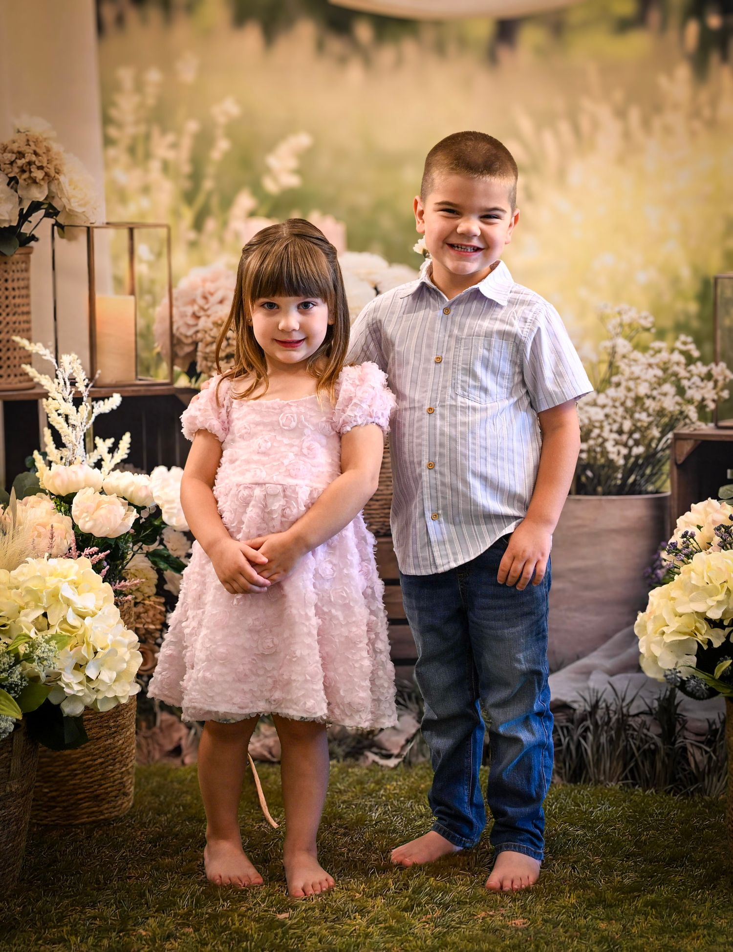 Two young children, a girl in a pink dress and a boy in a blue shirt and jeans, pose together among white flowers.