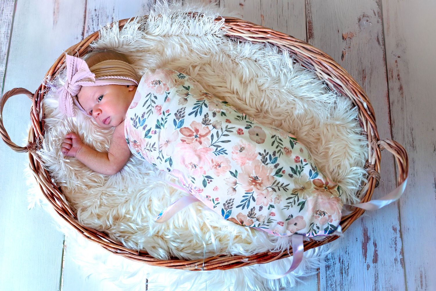 Peaceful newborn baby girl in floral swaddle with pink bow headband sleeping in wicker basket with fluffy lining.