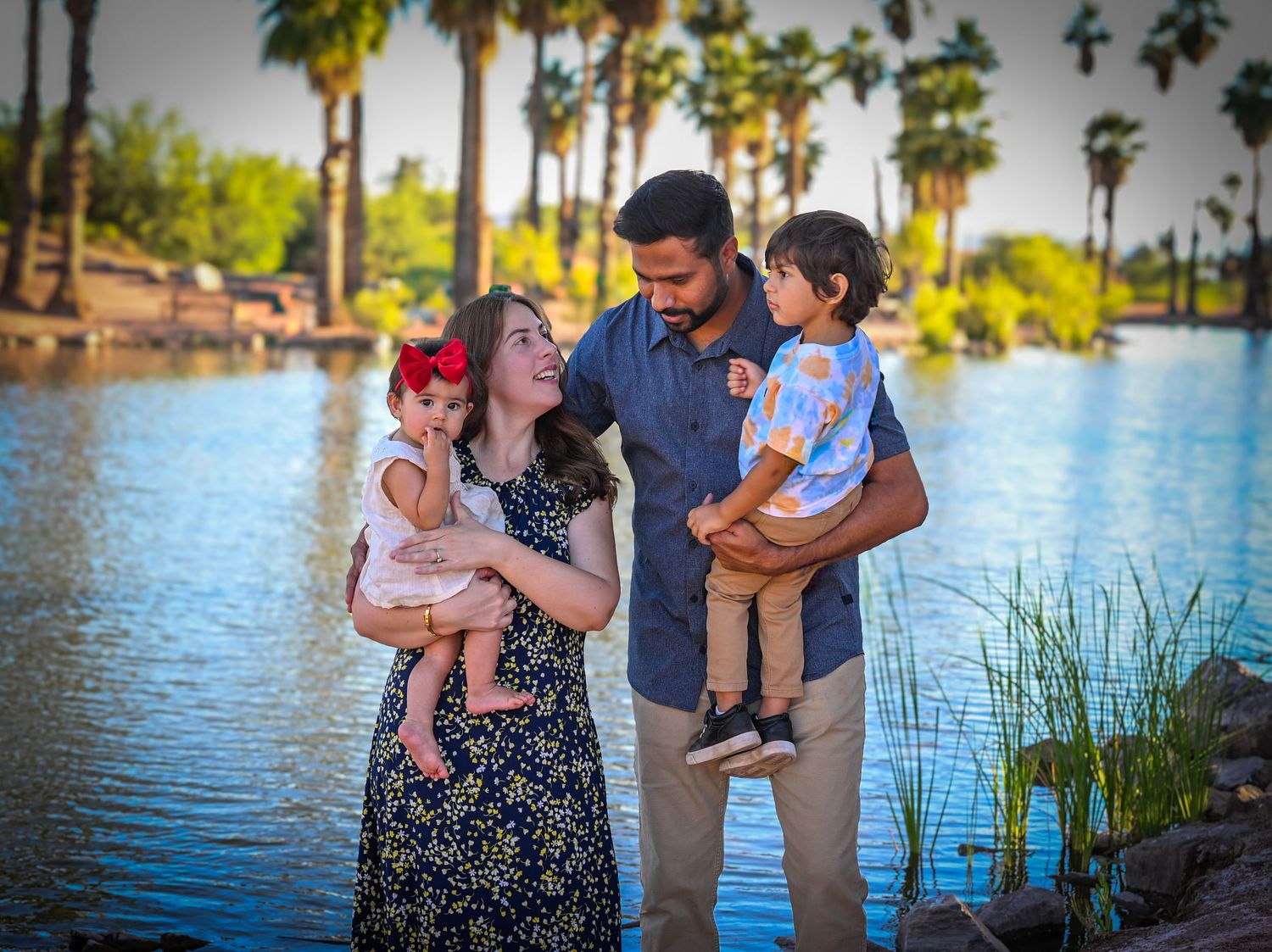 Family of four posing by a serene lake surrounded by palm trees, parents holding two young children outdoors.