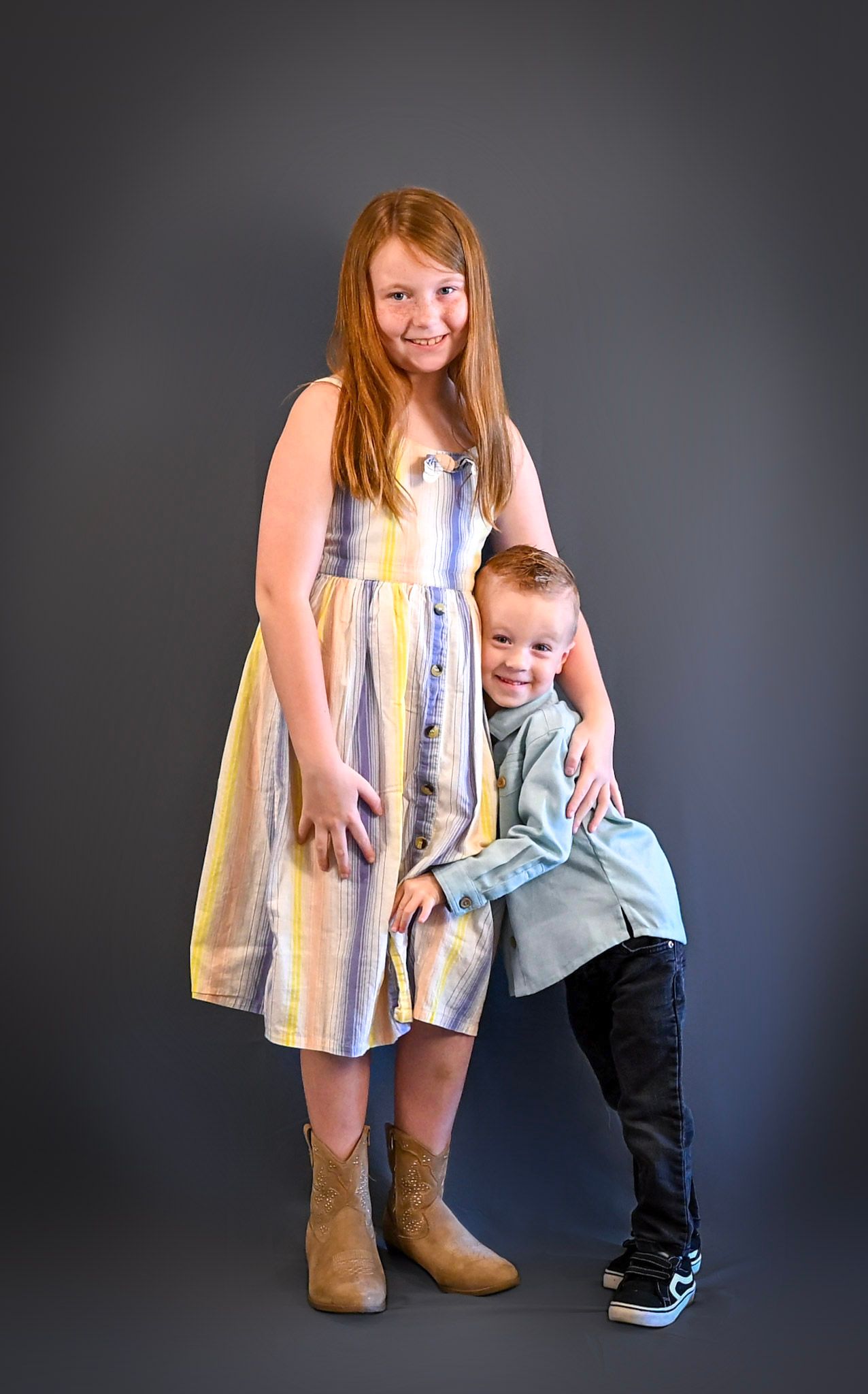 Two children stand together against a gray background wearing casual clothing including a striped dress and boots.