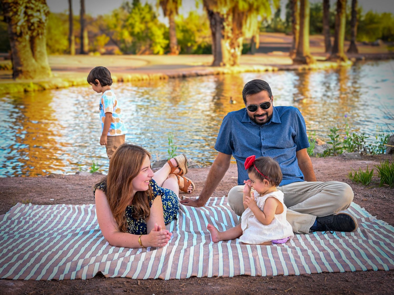Family enjoying a sunny picnic by a tranquil pond, with two adults and two young children on a striped blanket.