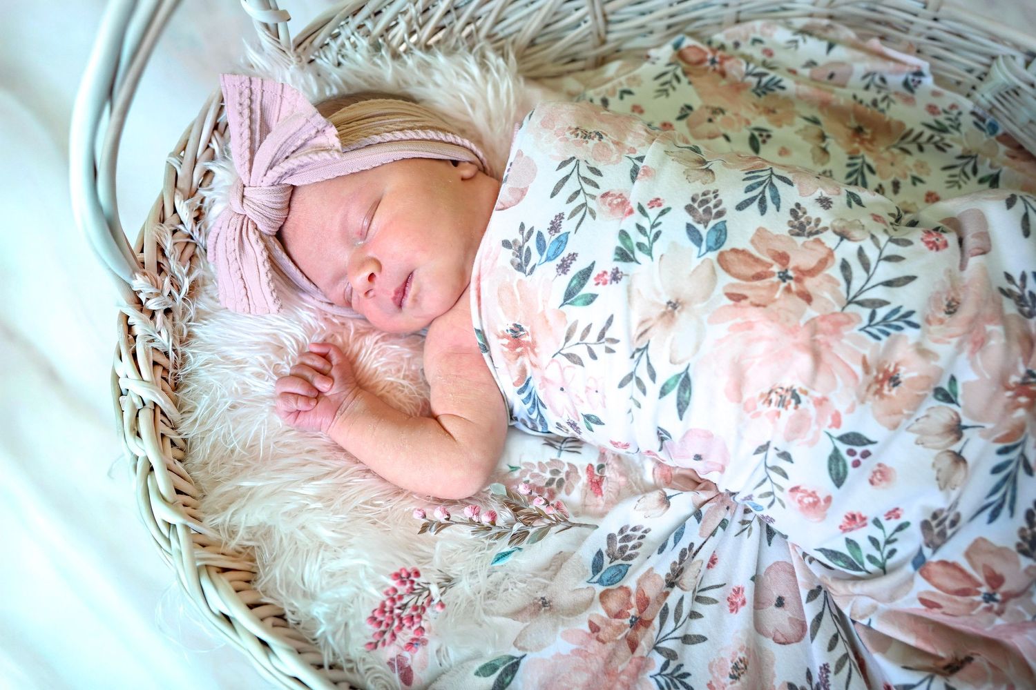 Sleeping newborn baby girl in floral swaddle with pink headband in wicker basket.