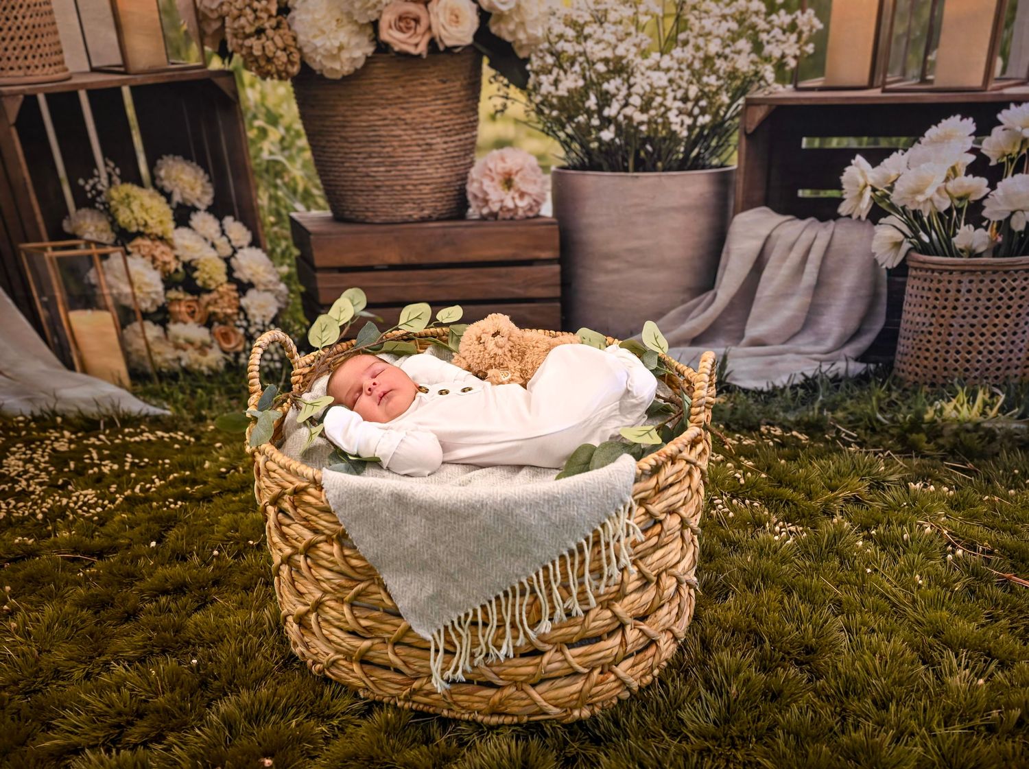 Newborn baby sleeping peacefully in a woven basket with white wrap and greenery on grass in a floral studio setting.