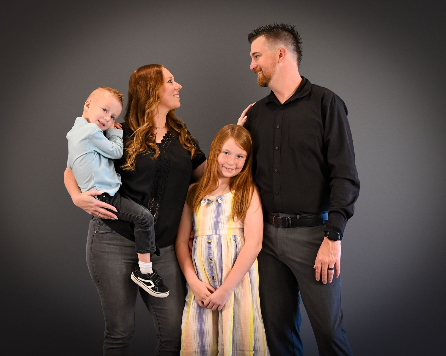 Family of four posing for a studio portrait, parents with red and dark hair holding a baby and standing with a young girl.