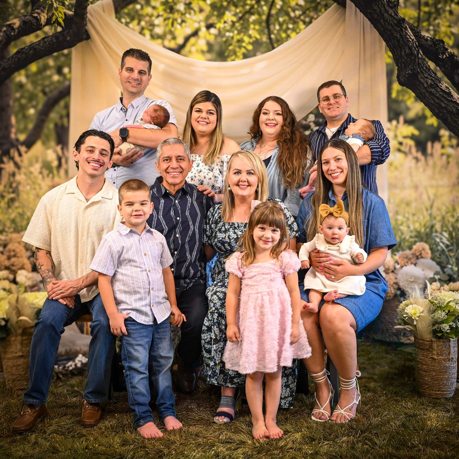 Large multigenerational family portrait outdoors with adults and young children posing together in a lush garden setting.