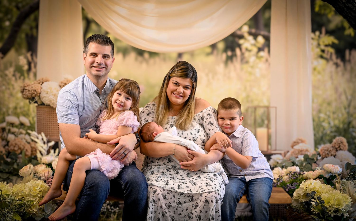 Happy family of five posing outdoors with newborn baby, two young children, and parents in a floral garden setting.