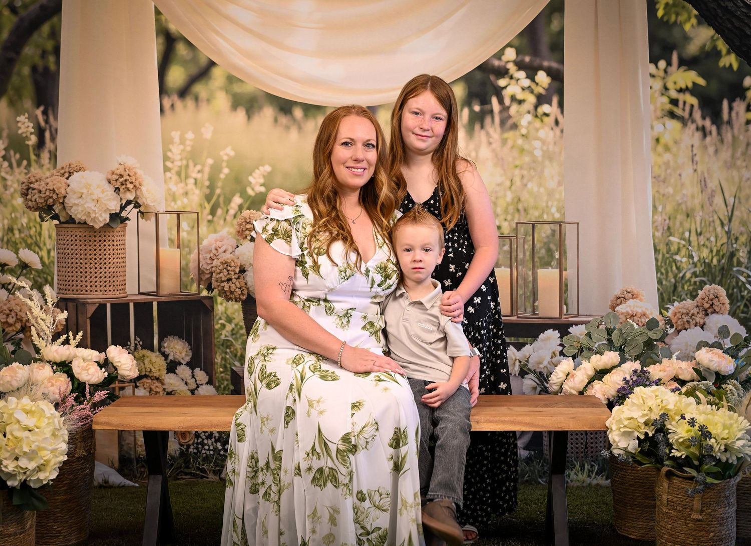 A smiling redheaded mother poses with her two children in front of a beautiful floral backdrop at an outdoor photo shoot.